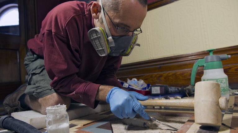 Man kneeling on the floor in the Sunnycroft entrance hall while working on the tiles with a hand tool